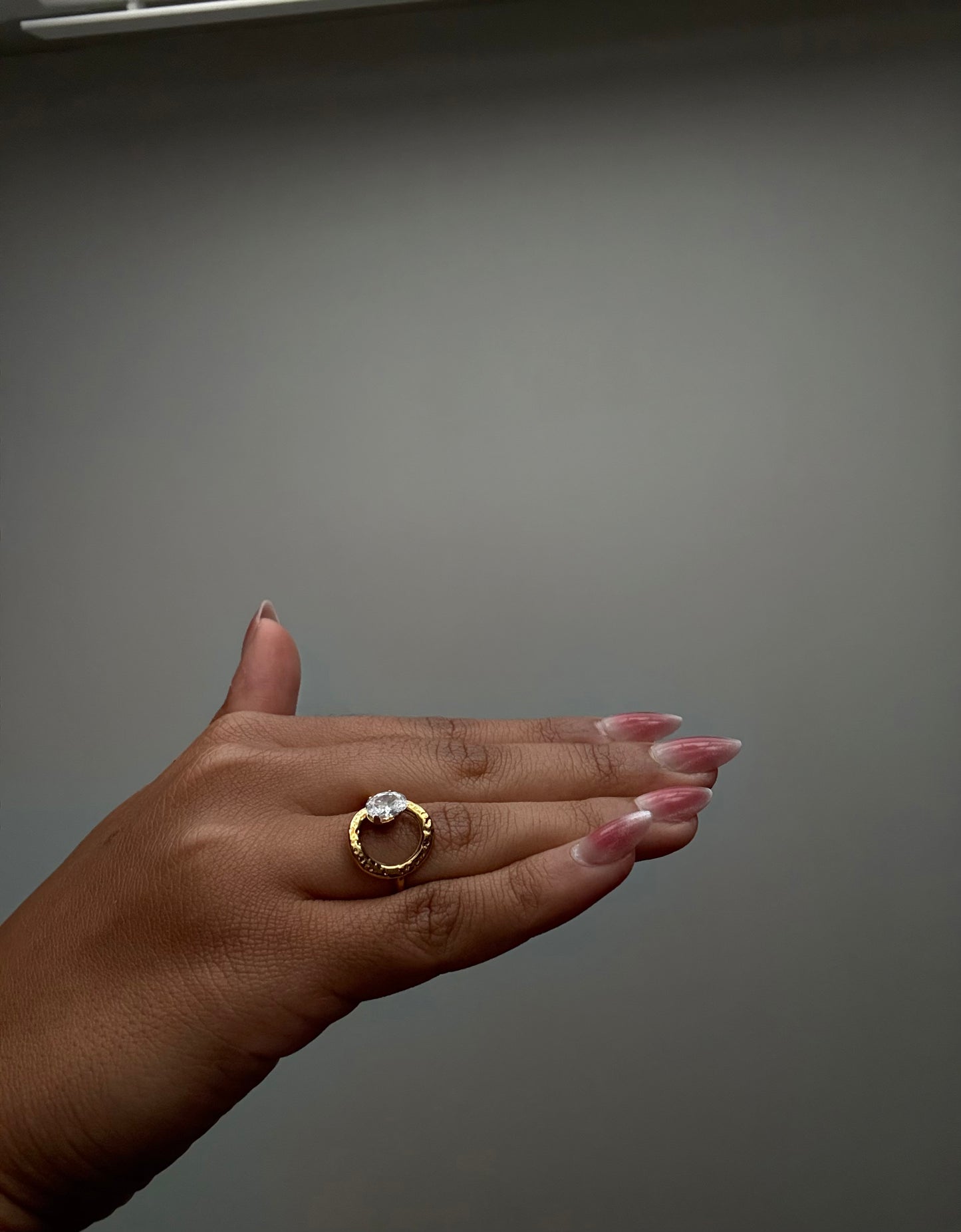 Hand wearing a gold ring with a clear gemstone against a gray background