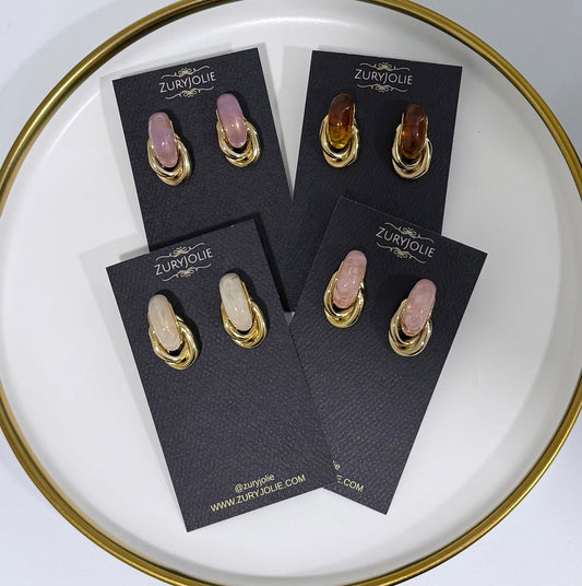 Four pairs of gold hoop earrings with oval cabochon gemstones in pink, peach, and cream tones, displayed on black ZURYJolie brand cards on a white plate with a gold rim.