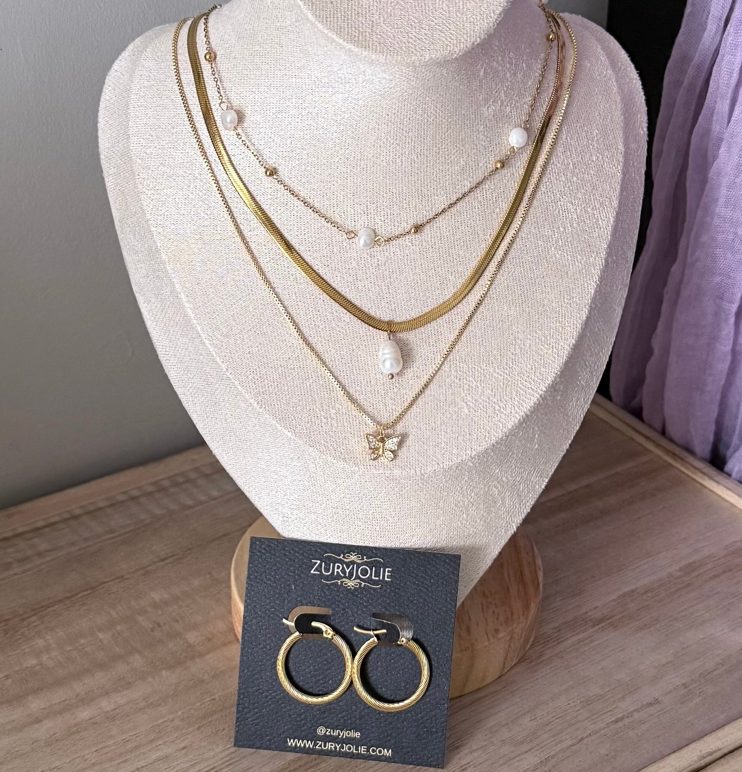 Mannequin bust with layered gold necklaces and a card displaying gold hoop earrings on a wooden surface.