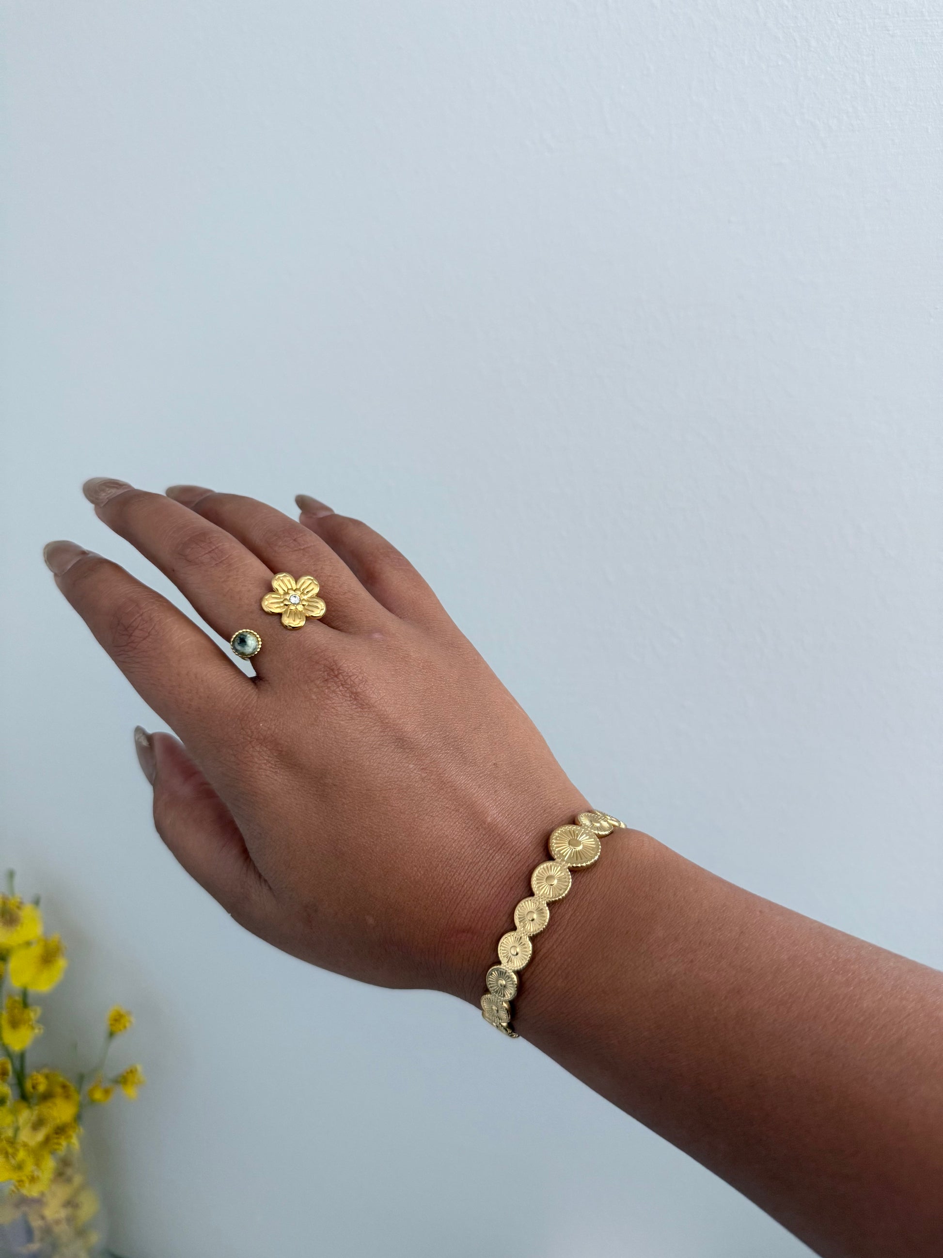Hand wearing a gold ring and bracelet on a light gray background with yellow flowers.