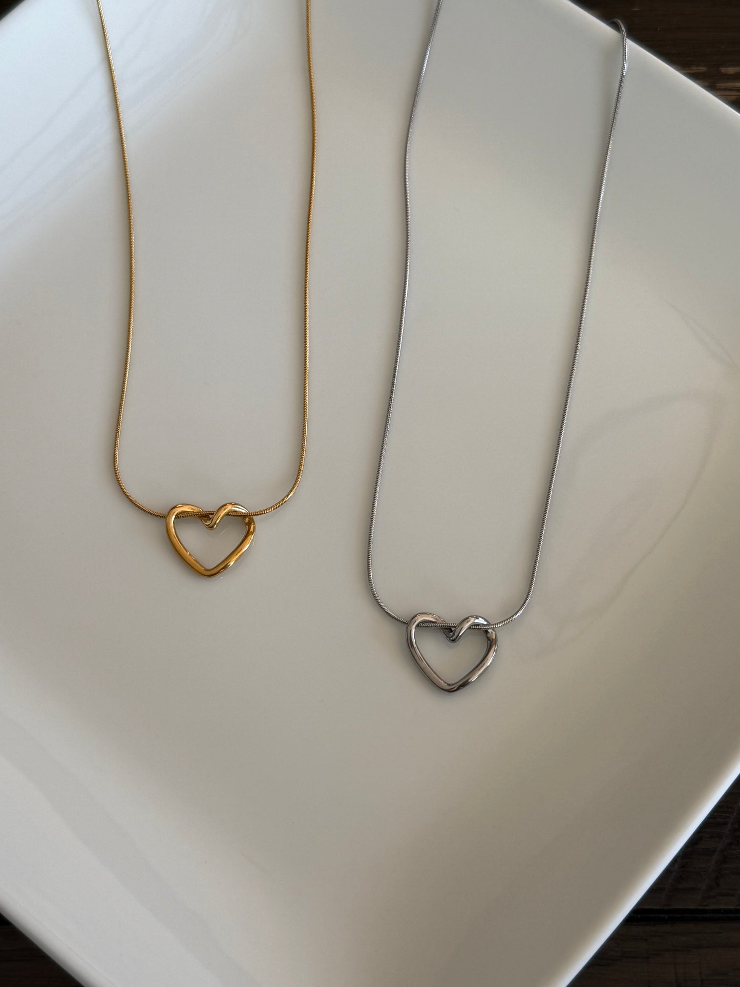 A minimalist necklace display featuring two heart-shaped pendant necklaces—one in gold-plated stainless steel and the other in silver stainless steel