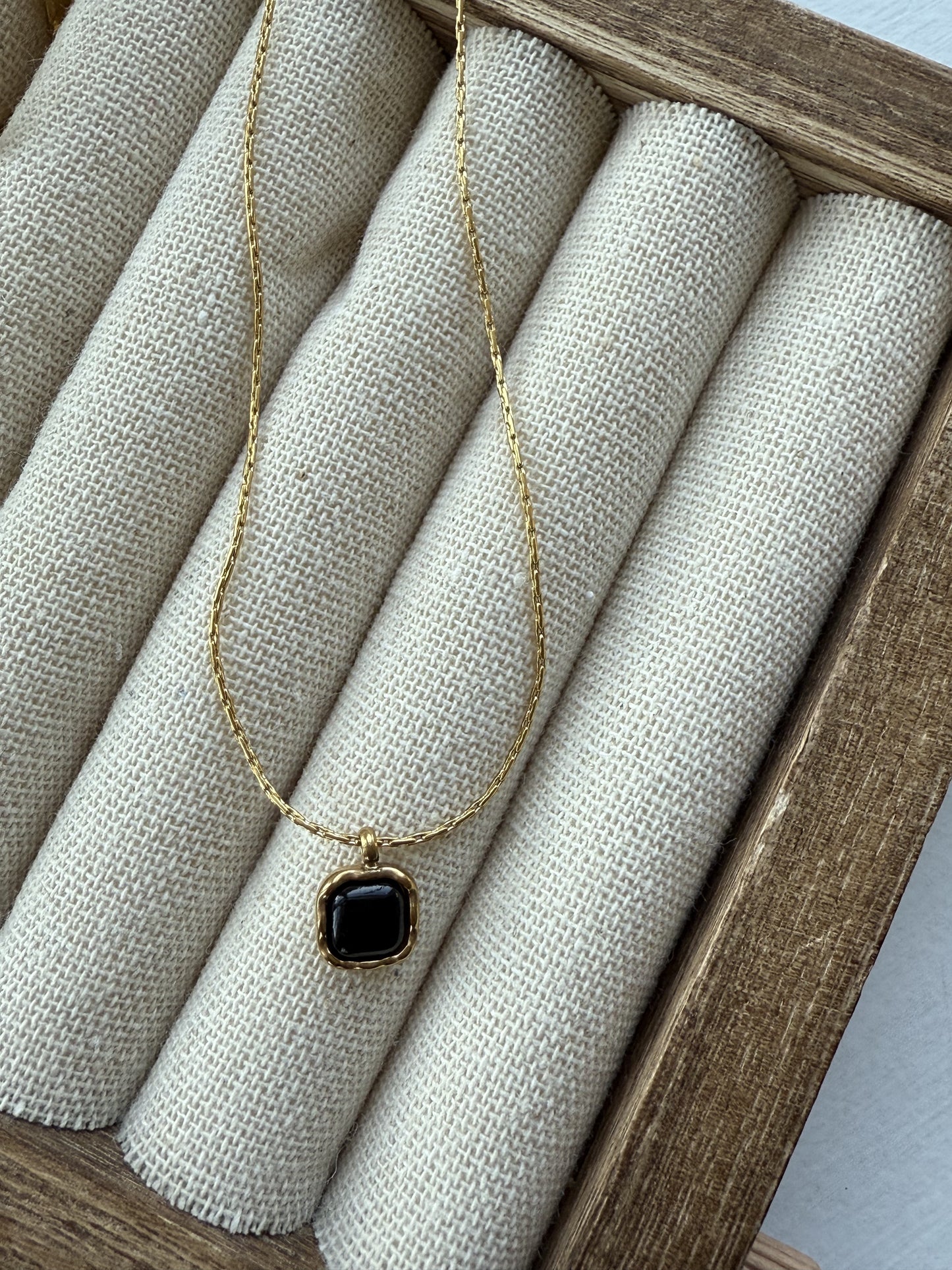 A gold-plated necklace featuring a delicate chain and a black square-shaped pendant with a wavy, organic border