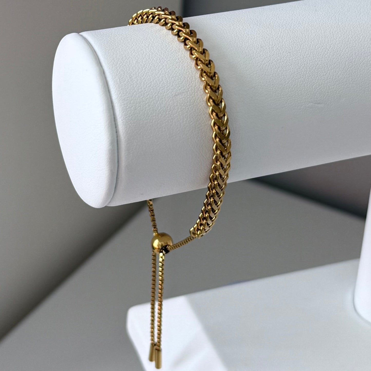 Gold chain bracelet on a white cylindrical stand against a neutral background