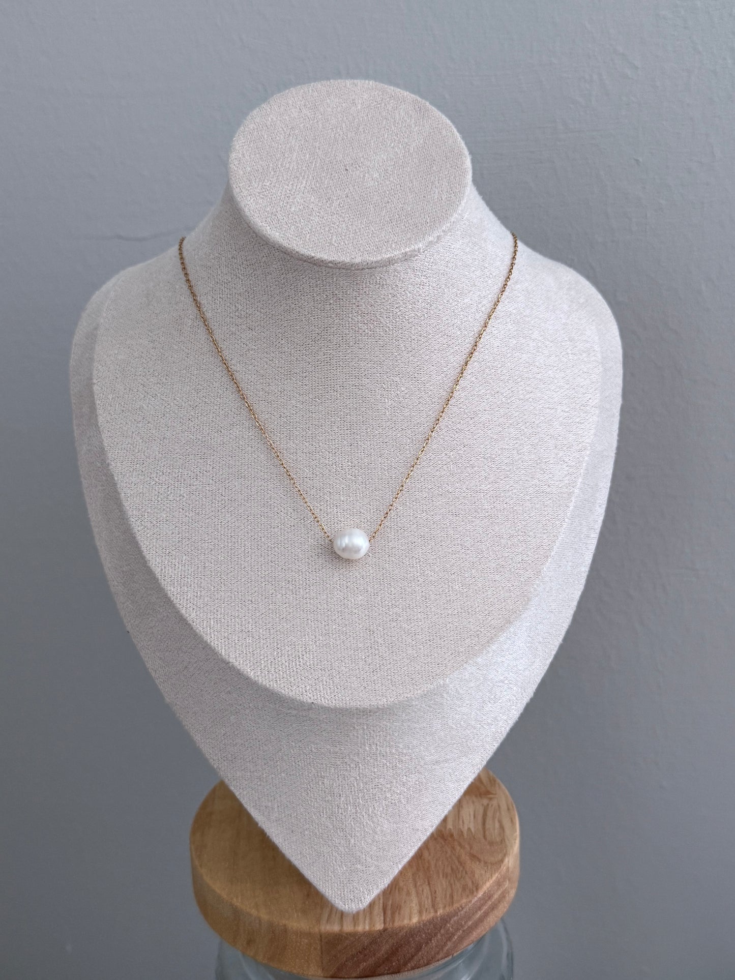 A delicate gold chain necklace with a single white pearl pendant, displayed on a textured cream-colored jewelry bust against a gray background.