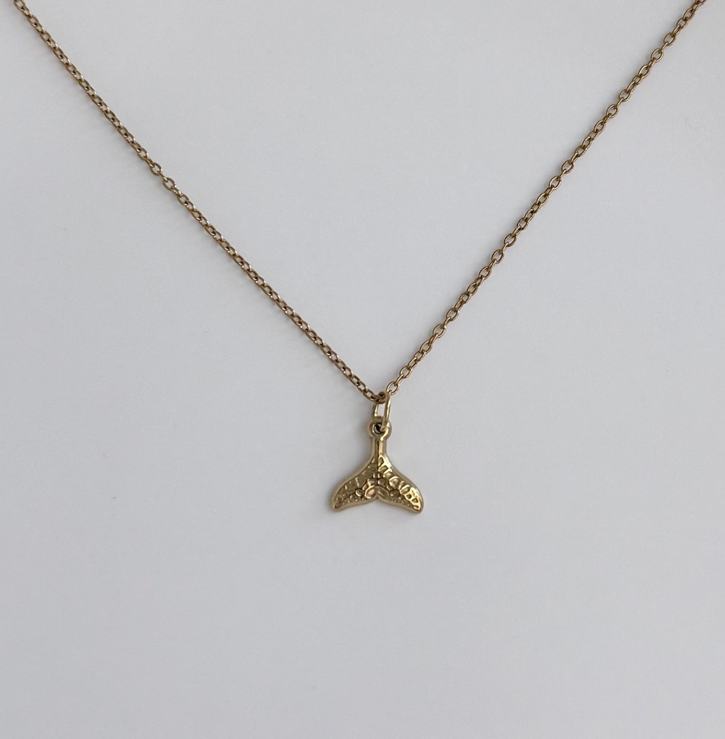 A gold chain necklace with a small textured gold whale tail pendant, displayed against a clean white background.