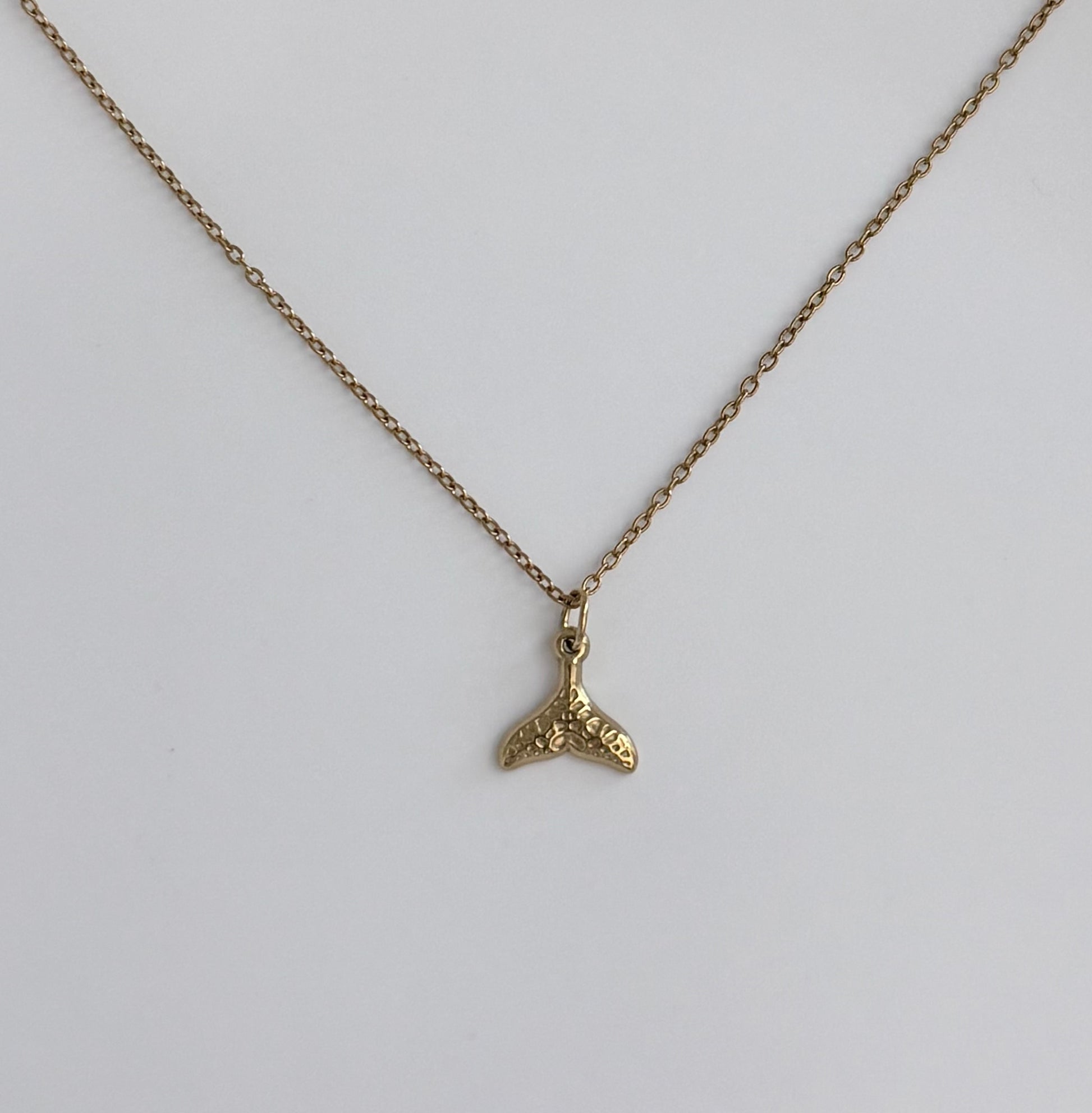 A gold chain necklace with a small textured gold whale tail pendant, displayed against a clean white background.