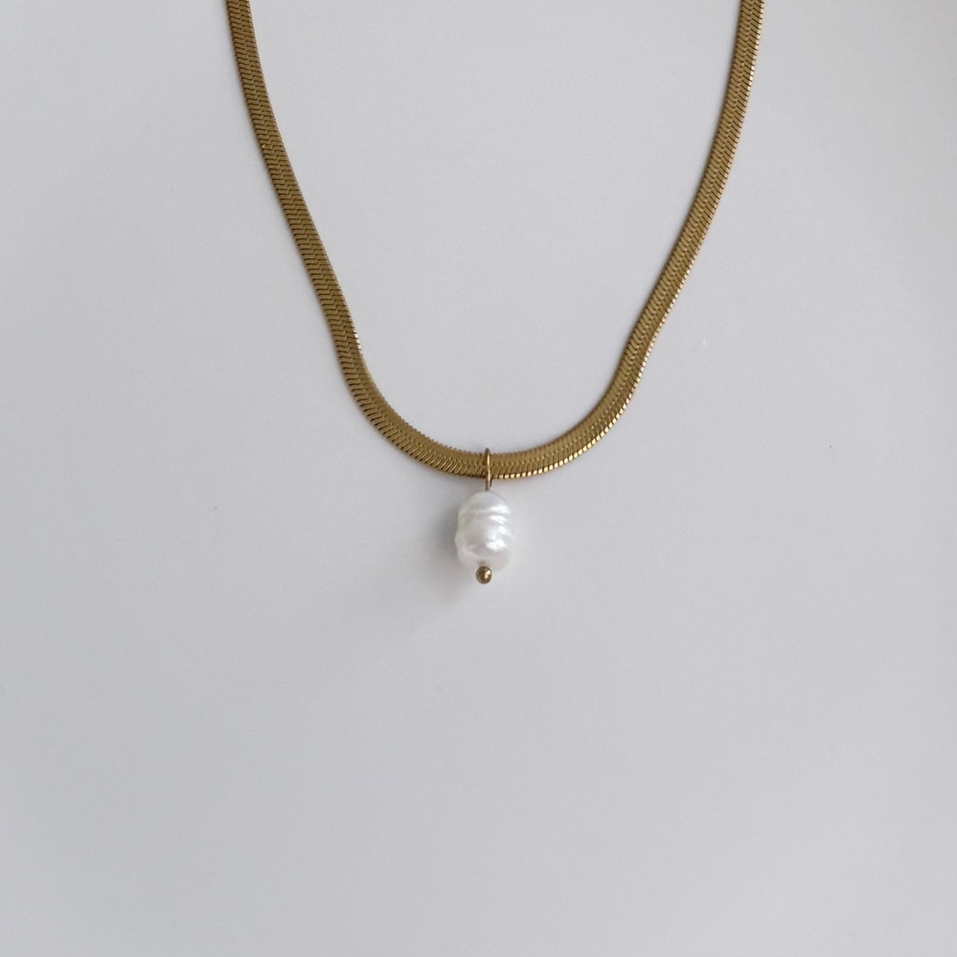 A gold snake chain necklace with a single white baroque pearl pendant, photographed against a plain light gray background.