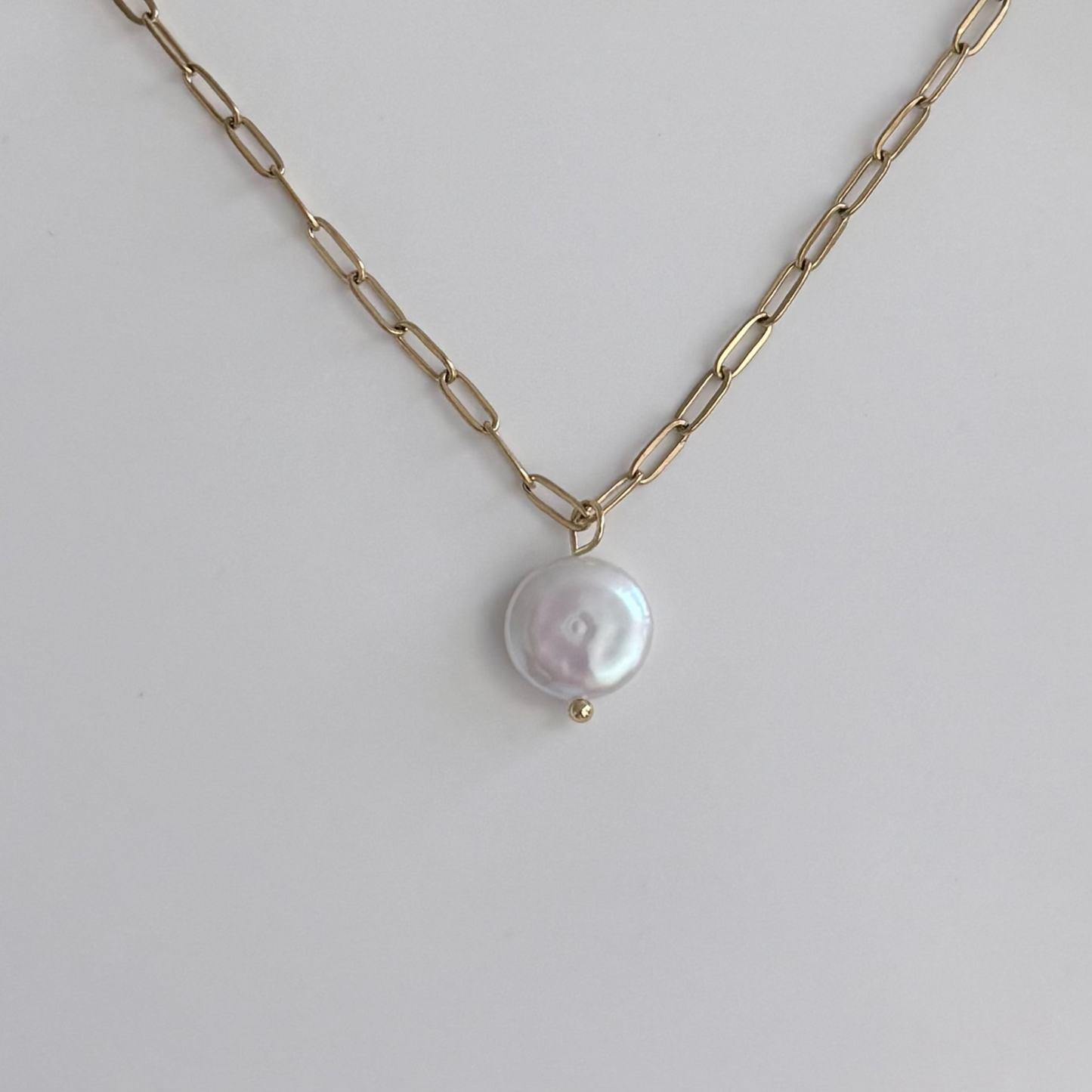 A gold paperclip chain necklace with a single round, lustrous white baroque pearl pendant, displayed on a clean light gray background.