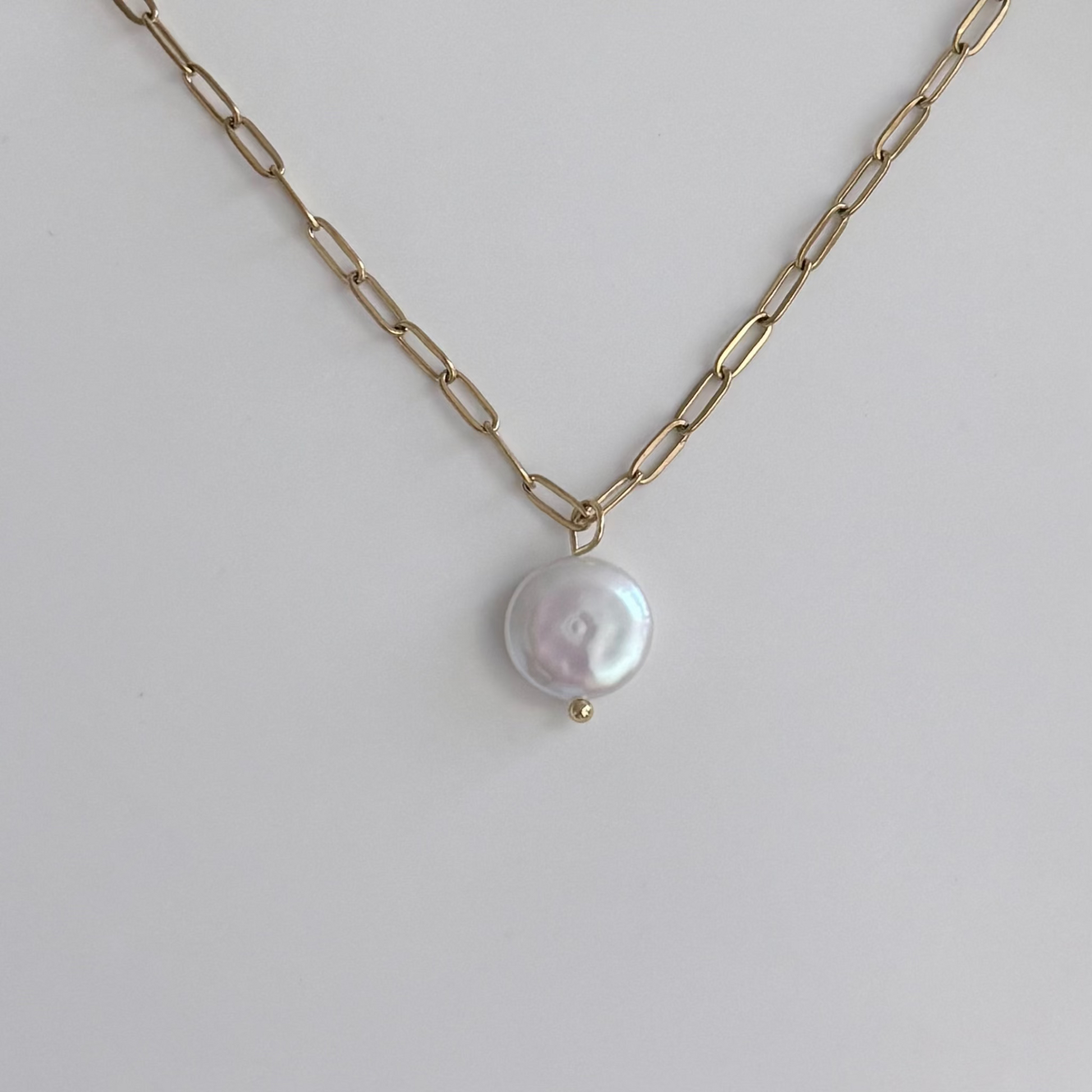 A gold paperclip chain necklace with a single round, lustrous white baroque pearl pendant, displayed on a clean light gray background.