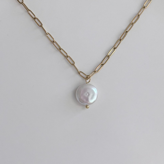 A gold paperclip chain necklace with a single round, lustrous white baroque pearl pendant, displayed on a clean light gray background.