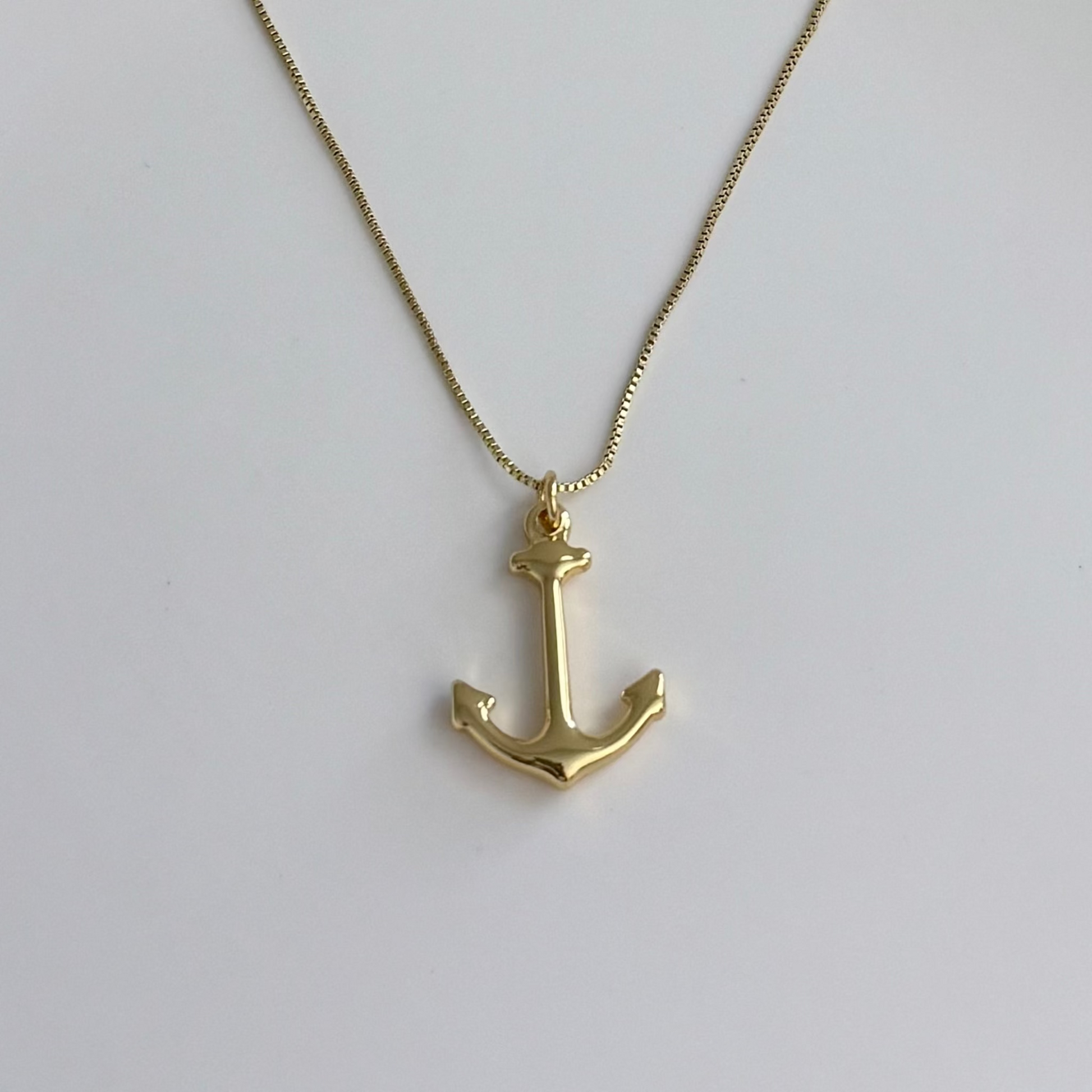 A gold anchor pendant necklace on a delicate gold chain, displayed against a clean, light gray background.