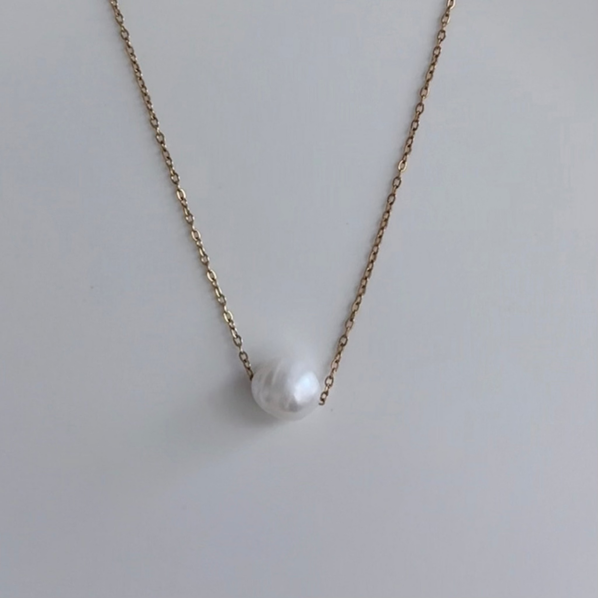 A delicate gold chain necklace featuring a single, irregular white baroque pearl pendant, displayed against a clean, light gray background.