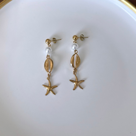 A pair of gold-toned dangle earrings featuring a small white pearl, a textured shell charm, and a dangling starfish, displayed on a white surface.